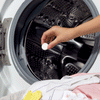 A hand places a white cleaning tablet inside an open washing machine drum. The machine is metallic, with a pile of clothes, including a yellow sock, visible in the foreground.