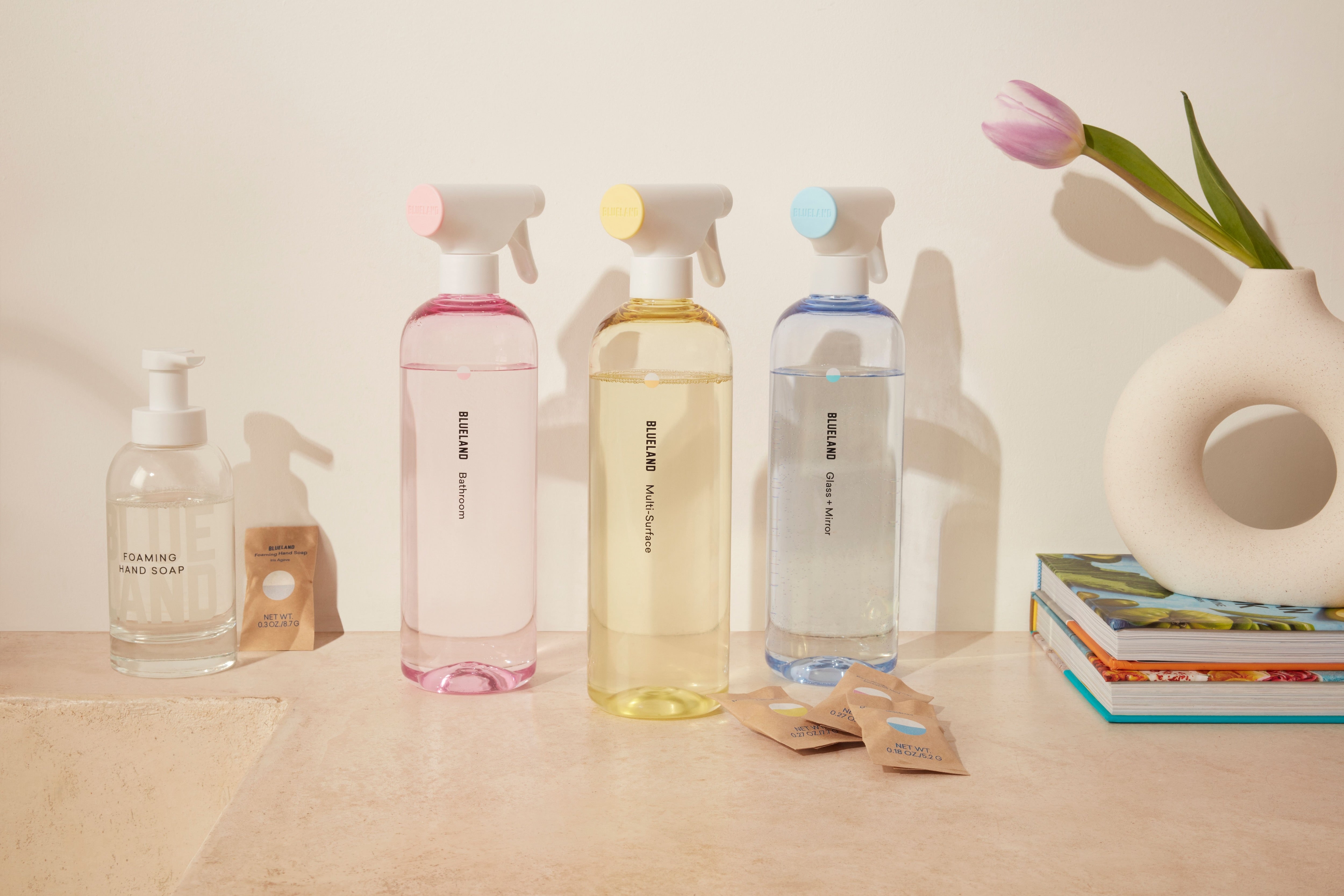 Four cleaning spray bottles and a liquid soap dispenser are arranged on a light countertop next to a modern white vase with a pink tulip and a small stack of books. Cleaning refill packets lie nearby.