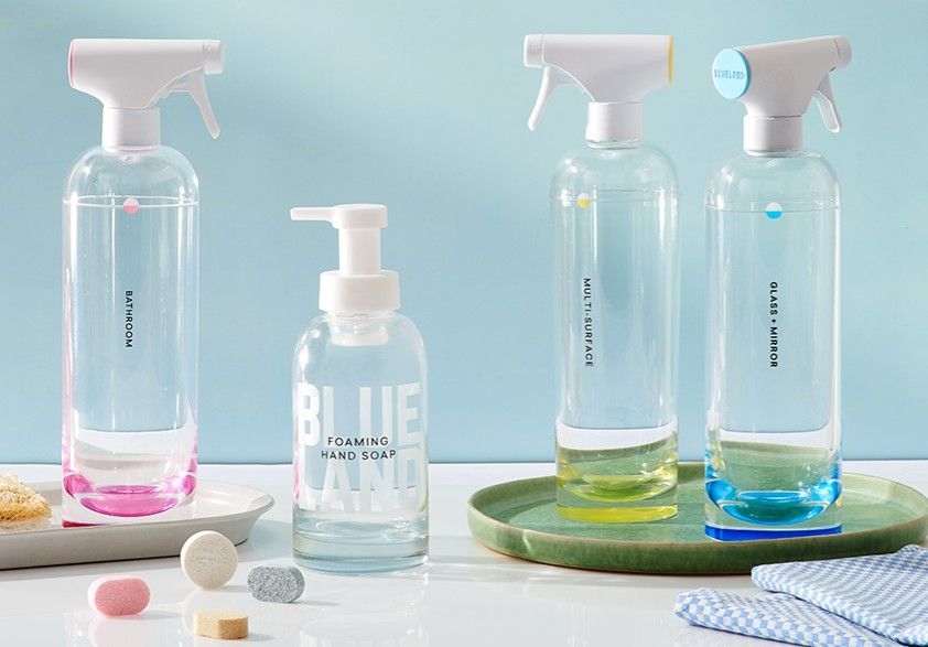 Four clear bottles labeled Bathroom, Multi-Surface, Glass & Mirror, and Foaming Hand Soap are arranged on a counter, each with a colored liquid and a matching tablet, with a light blue background.