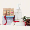 The Holiday Hand Soap Kit features a clear foaming dispenser, refill tablets in brown packets, a silver tin, and festive accents like red ribbon, pinecone, and green foliage, all arranged on a white round base against a light background.