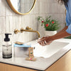 A person washes their hands with foaming soap at a bathroom sink with a modern gold faucet. On the counter sits the Holiday Hand Soap Kit, along with a potted plant and a round mirror on a tiled wall.