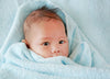 A baby wrapped in a soft blue blanket looks ahead with wide eyes. The infants head is covered with fine hair, and the blanket is snugly wrapped around their shoulders, creating a cozy and serene scene.