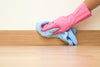 Person wearing pink rubber gloves cleaning baseboards with a blue cloth on a wood floor