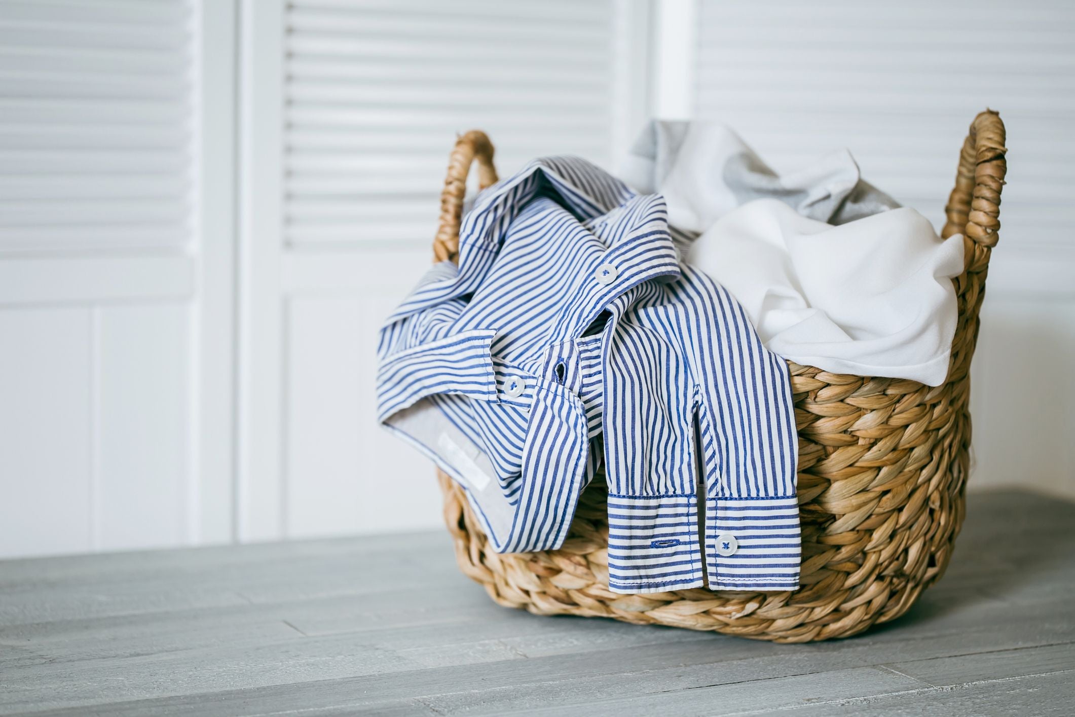 A woven basket filled with neatly folded laundry, including a blue and white striped shirt, sits on a light gray wooden surface in front of white louvered doors.