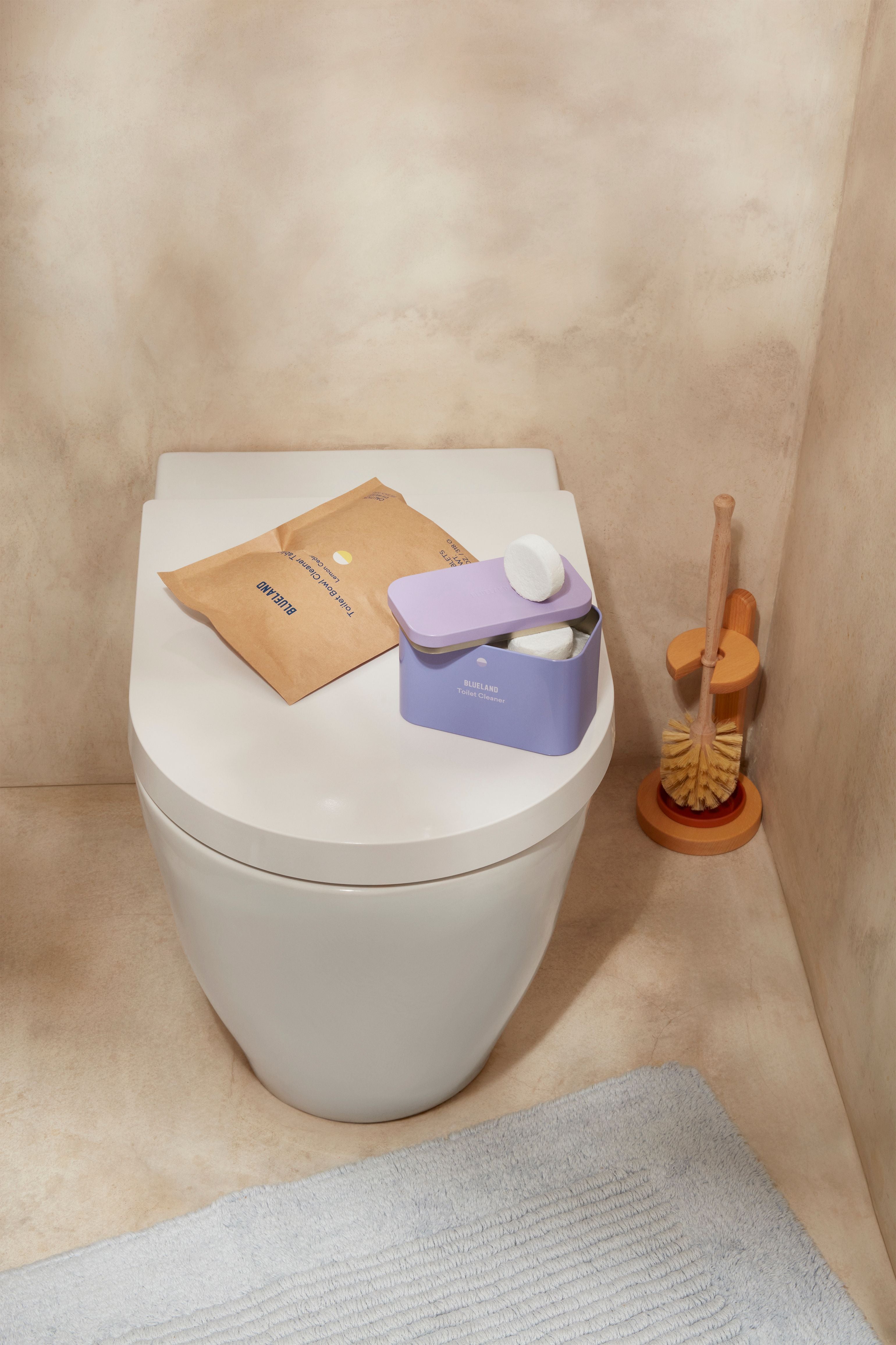 A modern white toilet with a lavender container, brown paper package, and white cleaning tablets on top. A toilet brush in an orange holder stands nearby on a beige floor with a blue rug.