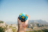 A hand holding a small globe against a background of scenic mountains and a clear blue sky. The globe shows parts of North and South America. The scene conveys a sense of exploration and global unity.