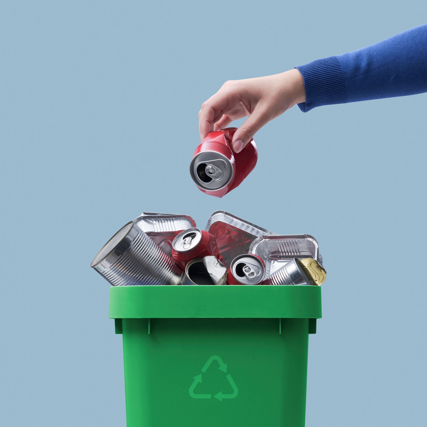 Hand placing a red can into a green recycling bin with a blue background