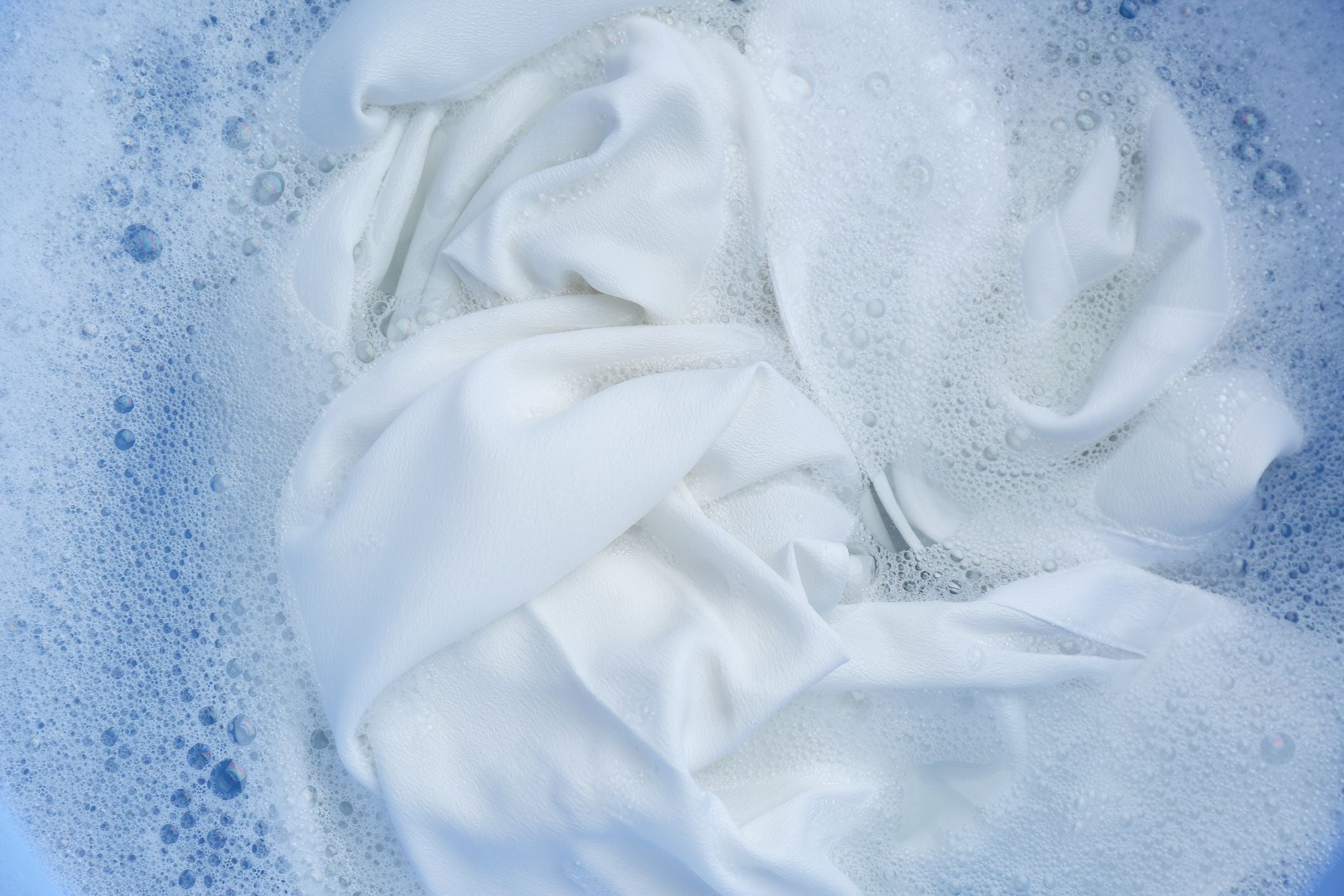White fabric soaking in soapy water, with bubbles and foam visible on the water’s surface, suggesting the process of washing or cleaning laundry.