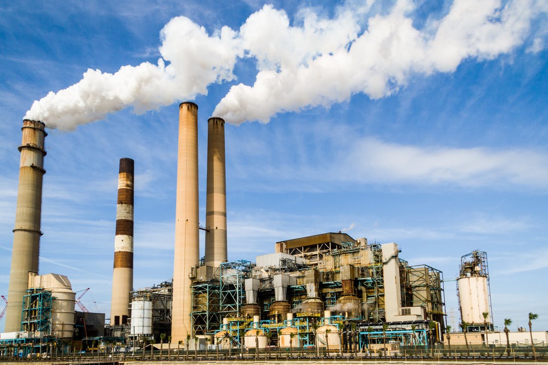 A large industrial power plant with several tall smokestacks emitting white smoke into a blue sky, surrounded by various pipes and metal structures.