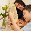A child holding a spray cleaner bottle at the sink with his smiling mom in the background
