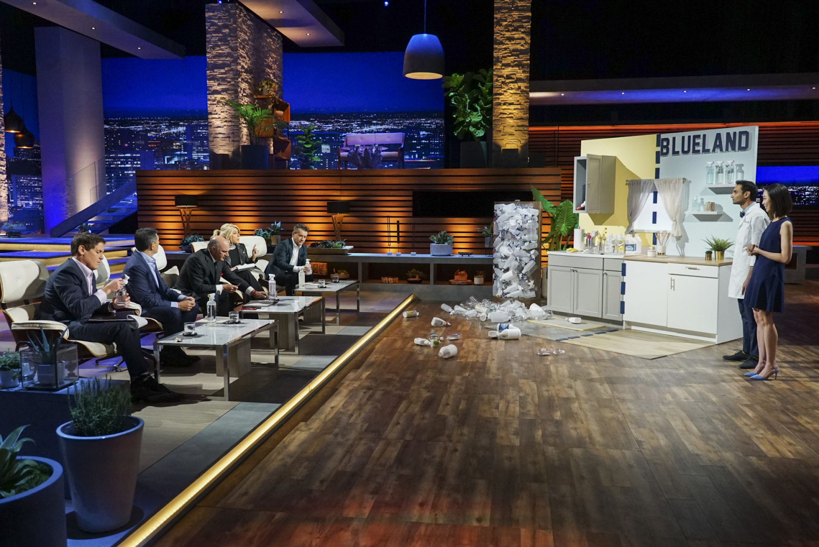 Two entrepreneurs in lab coats present a product called Blueland to a panel of five seated investors on a brightly lit stage, with plastic waste and a modern kitchen setup displayed beside them.