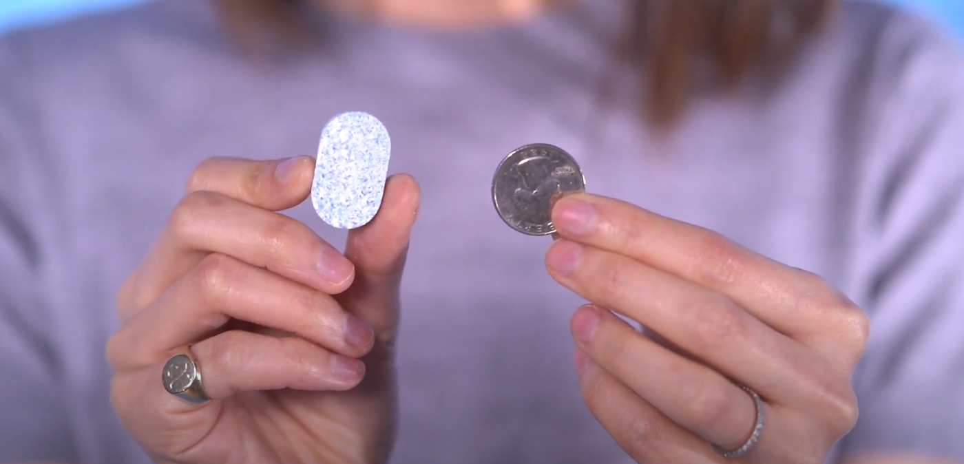 A person holds a speckled, oval-shaped tablet in one hand and a quarter coin in the other, with both objects held up for size comparison.
